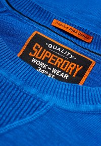 Blauwe katoenen sweatshirt met ribgebreide halslijn en label met "SUPERDRY" in oranje op een zwarte achtergrond, wat aangeeft dat het onderdeel is van de werk-kledingcollectie.