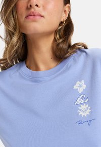 Jasnoniebieska bawełniana koszulka z krótkim rękawem, ozdobiona kwiatowymi i falistymi grafikami w kolorze białym i niebieskim. Okrągły dekolt i krótkie rękawy z widocznym przeszyciem.