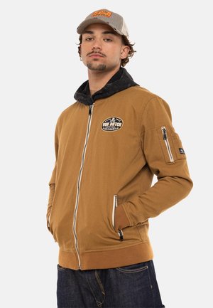 Homme portant une veste marron zippée avec un logo écusson sur la poitrine et une casquette beige rayée, les mains dans les poches, faisant face à l'avant avec une expression neutre.