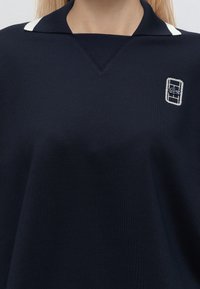 Personne portant une chemise à col bleu marine avec des accents blancs et un petit logo rectangulaire brodé sur la poitrine gauche.