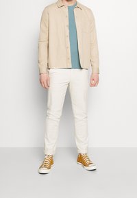 Béžová košile s dlouhým rukávem přes tyrkysové triko, doplněná bílými kalhotami a béžovými teniskami. Košile má knoflíkový zapínání a kapsu.