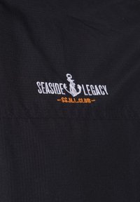 Zwarte stof met wit geborduurde anker tussen de woorden „Seaside Legacy“ en oranje tekst „S.S.N.L. Club“ daaronder.