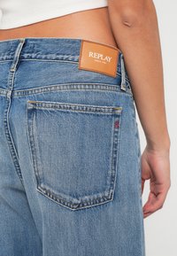 Ανοιχτού μπλε τζιν παντελόνι με υφή, που διαθέτει ένα καφέ δερμάτινο ετικέτα στο πίσω μέρος, ορατές ραφές και μια διακριτική κόκκινη λεπτομέρεια λογότυπου.