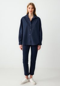 Navy blue button-up shirt with a pocket, paired with matching slim-fit pants and white sneakers. Smooth fabric with long sleeves.