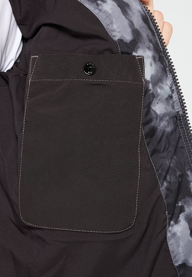 Czarna materiałowa kieszeń z zapięciem na guzik, umieszczona w kurtce z szaro-czarnym podszewką w stylu tie-dye. Widoczne detale przeszycia.