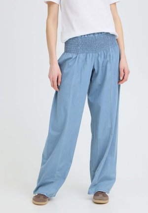 Osoba ubrana w jasnoniebieskie spodnie o szerokich nogawkach z marszczonym elastycznym pasem, białą koszulkę i brązowe trampki na jednolitym tle.