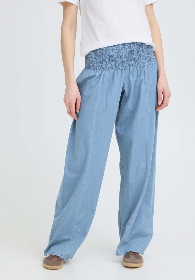 Personne portant un pantalon ample bleu clair à taille élastiquée smockée, un t-shirt blanc et des baskets marron sur un fond uni.