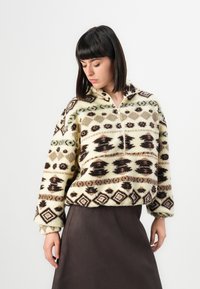 Vrouw met steil, donker haar die een fleecetop met een patroon en een halve rits draagt, samen met een donkere rok, tegen een effen achtergrond.