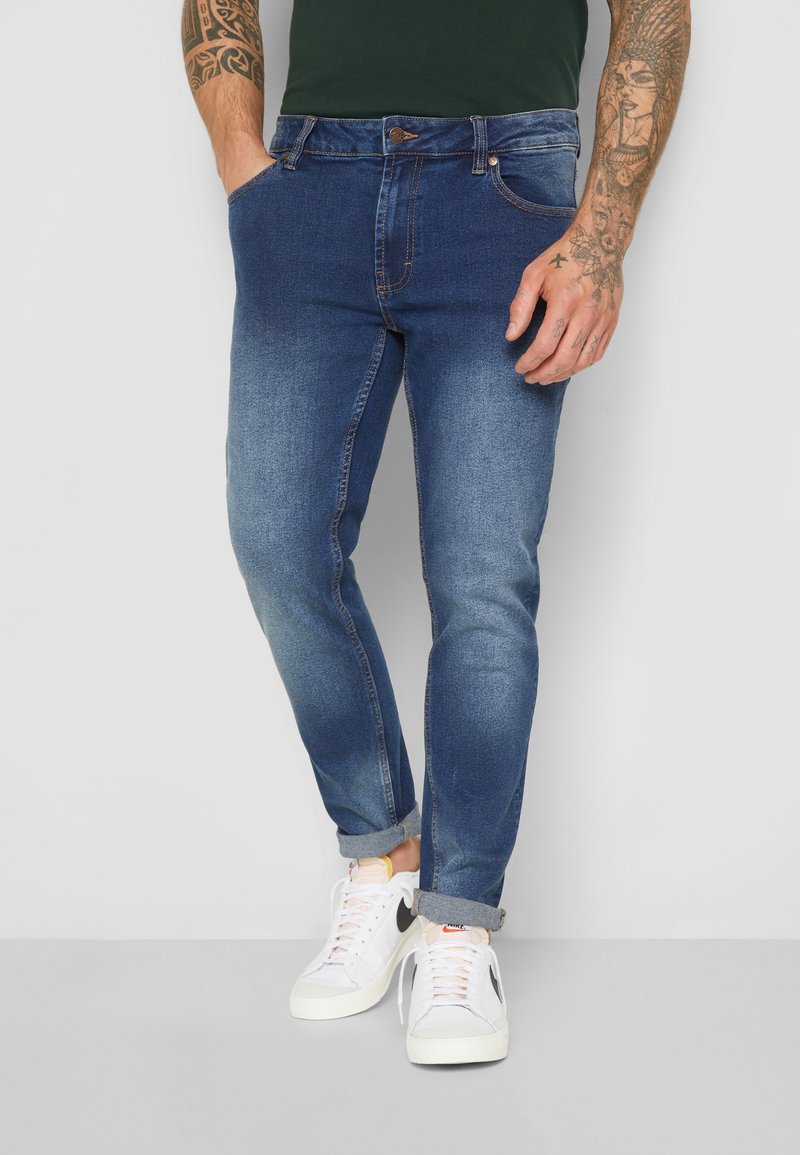 Fakó kék farmernadrág, slim fit szabással, elülső zsebekkel, gombos záródással és feltekert szárakkal, fehér sportcipőkkel párosítva.