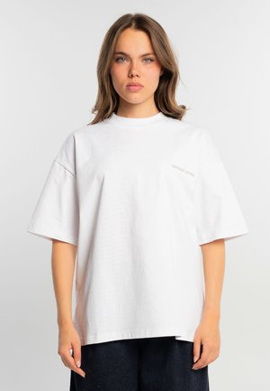 Giovane donna con capelli castani fino alle spalle, che indossa una maglietta bianca oversize e pantaloni scuri, in piedi contro uno sfondo chiaro e semplice.