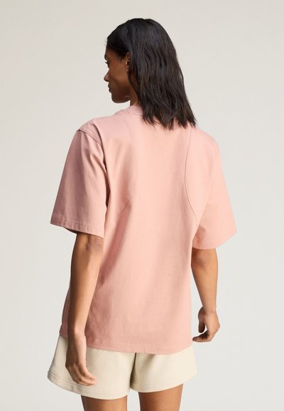Woman with shoulder-length black hair wearing a loose pale pink short-sleeve shirt and beige shorts, viewed from behind against a plain background.