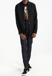 Una persona está de pie vistiendo una camisa negra de botones sobre una camiseta gráfica, combinada con jeans oscuros y zapatillas negras. El fondo es blanco liso.