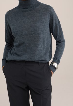Dark gray turtleneck sweater paired with black trousers, hands in pockets, and silver cuff bracelets on left wrist against a neutral background.