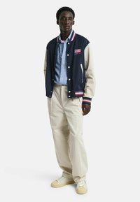 Giacca varsity blu navy e crema con bottoni bianchi, polsini a righe con coste, e dettaglio della bandiera USA, abbinata a una camicia a righe blu e pantaloni crema.