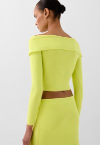 Ein amarilli-grünes Strickoberteil mit schulterfreiem Design und langen Ärmeln, kombiniert mit einem passenden Rock, der aus einem strukturierten Stoff mit subtilen Akzentnähte besteht.