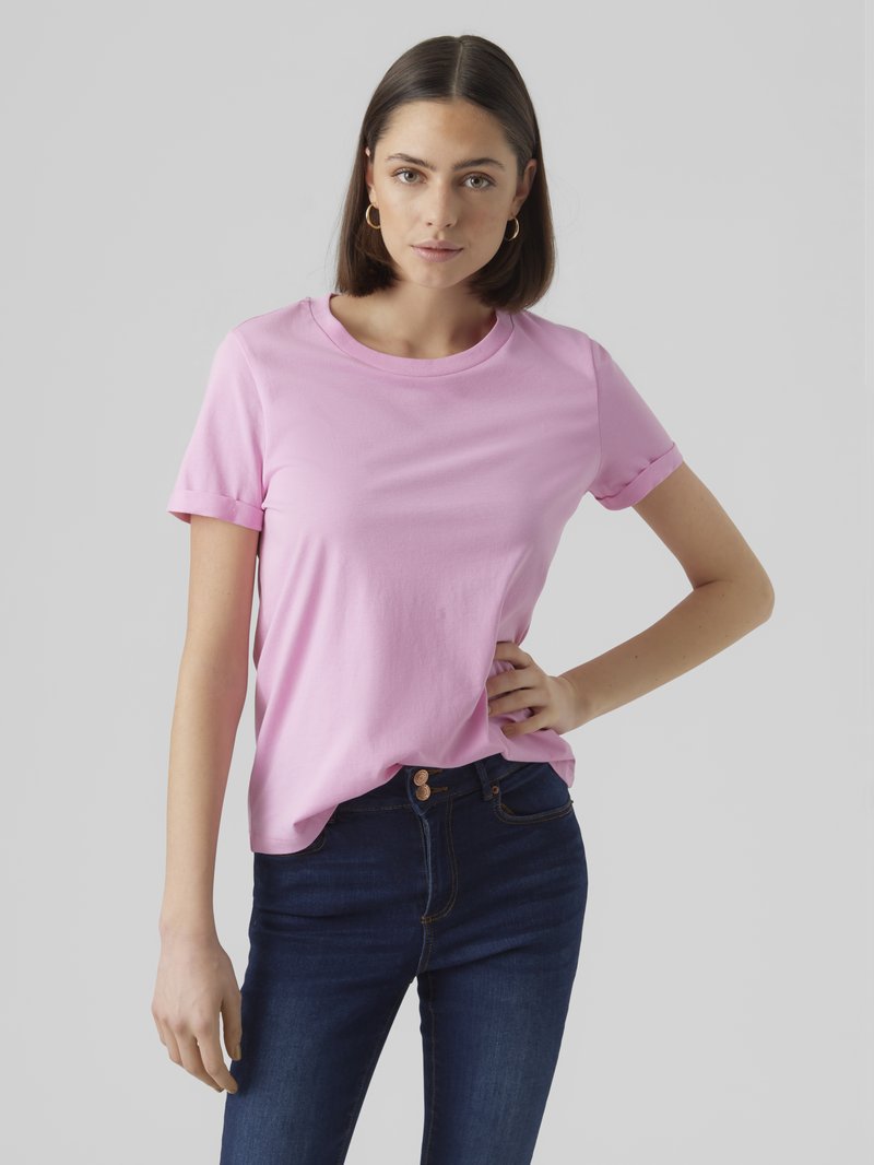 Ung kvinna med rakt brunt hår, bärande guldfärgade ringörhängen, en rosa kortärmad t-shirt och mörkblå jeans, stående med handen på höften.