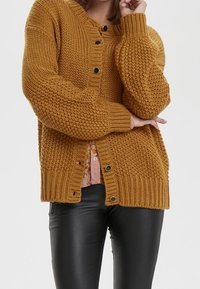 Sinapinkeltainen neulecardigan, jossa on tekstuuri kuvio, pyöreä kaula-aukko ja mustat napit, yhdistettynä vaaleanpunaiseen paitaan alla.