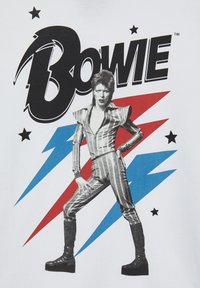 T-shirt bianco con grafica di una figura in un outfit a strisce metalliche con stivali neri, circondata da fulmini rossi e blu e stelle.
