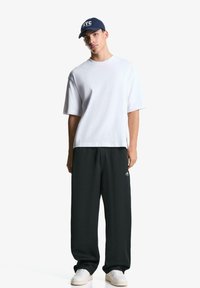 Ung mand står iført en hvid oversized T-shirt, sorte brede bukser, hvide sneakers og en marineblå kasket med NYC-tekst.