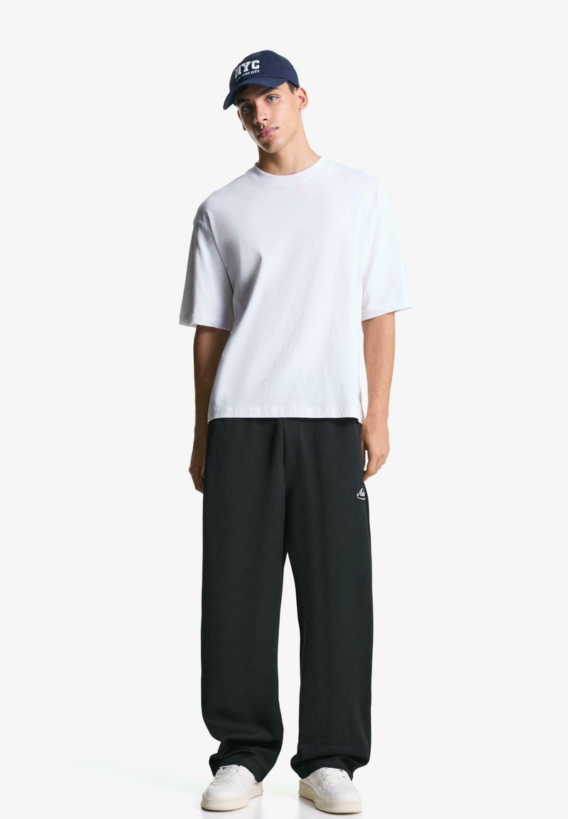 Ung mand står iført en hvid oversized T-shirt, sorte brede bukser, hvide sneakers og en marineblå kasket med NYC-tekst.