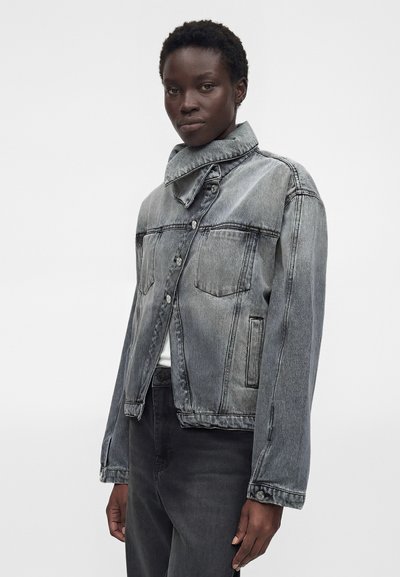 Μαύρο άτομο που φοράει ξεθωριασμένο γκρι τζιν μπουφάν με ψηλό, ασύμμετρο γιακά με κουμπιά και μαύρο παντελόνι, στέκεται μπροστά σε απλό λευκό φόντο.