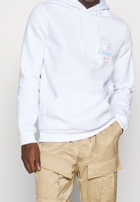 Felpa bianca con cappuccio, dotata di tasca frontale a marsupio e grafica testuale in blu e rosa sul petto, indossata su pantaloni cargo beige.