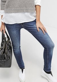 Kvinna som bär blå smala jeans, vita slip-on skor, grå stickad väst över vit skjorta och håller en svart handväska vid sin sida.