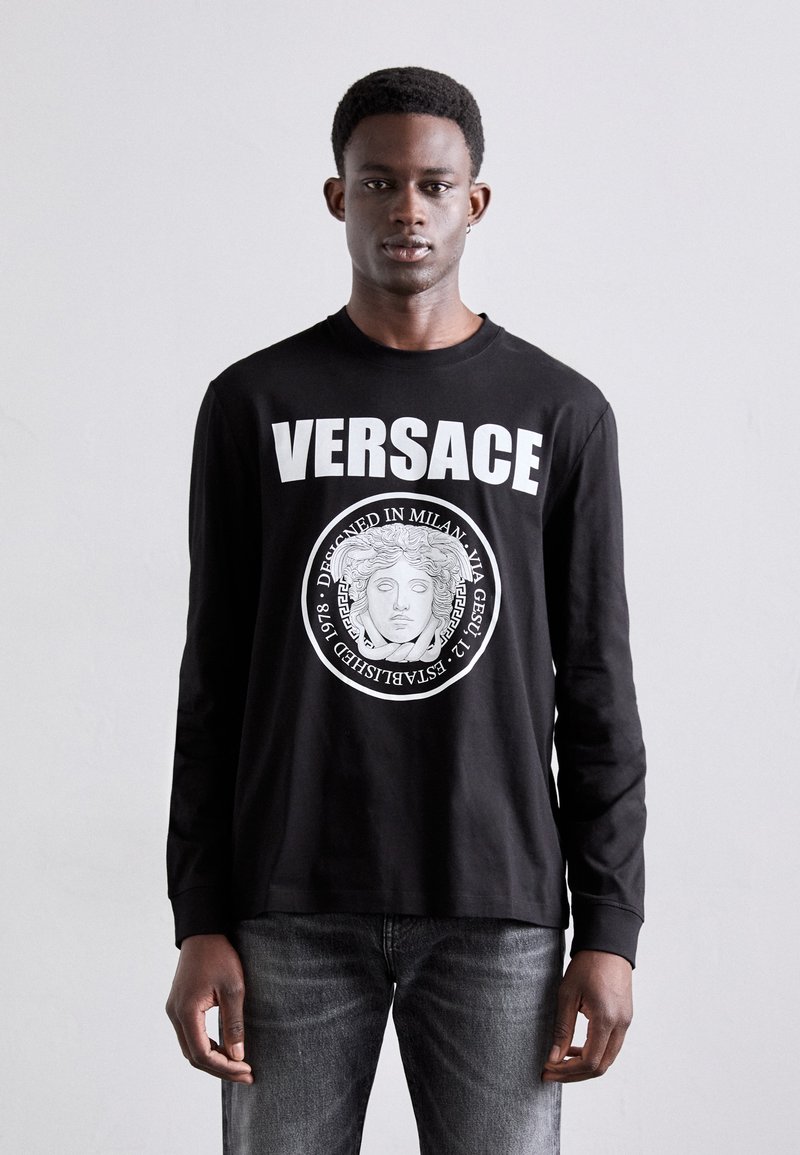 Fekete, hosszú ujjú pamut póló, fehér "VERSACE" felirattal és klasszikus maszk mintázatú kör alakú grafikával. Slim fit.