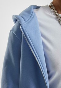 Felpa blu chiaro con zip e texture morbida, caratterizzata da un ampio cappuccio e una cerniera argentata. Indossata su una t-shirt bianca.