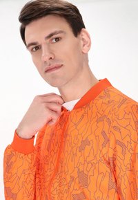 Giacca bomber arancione con un motivo geometrico testurizzato, dotata di zip frontale e polsini a costine. Tessuto leggero con una finitura liscia.