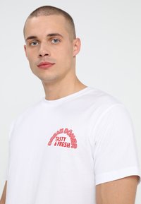 Bijela majica kratkih rukava s crvenim grafičkim otisakom koji glasi "DREAM DÖNER TASTY & FRESH." Glatka pamučna tekstura.