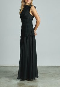 Schwarzes Abendkleid mit ärmellosem Design, plissierter Textur, tailliertem Oberteil und fließendem Rock. Verfügt über Raffungen und einen hohen Halsausschnitt.