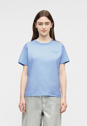 Jeune femme aux cheveux bruns lisses portant un T-shirt bleu clair à manches courtes et un pantalon ample argenté, debout devant un fond blanc.