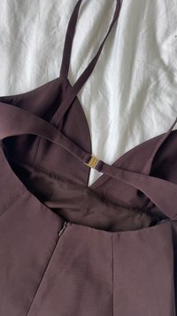 Hnedé šaty bez rukávov s priliehavým korzetom, prekríženými ramienkami a zlatými doplnkami. Obsahujú skrytý zips a hladkú textúru látky.