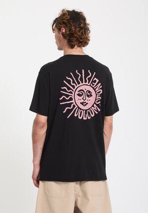 Homme aux cheveux bouclés portant un t-shirt noir avec un dessin de visage de soleil rose et le texte "Volcom" au dos, pantalon beige, dos tourné.