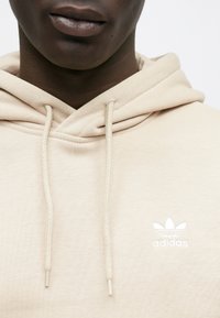 Bézs színű pamut kapucnis pulóver behúzózsinóros kapucnival, bal oldali mellrészen fehér hímzett Adidas logóval. Simább textúrájú, hétköznapi dizájn.