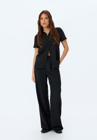Schwarz kurzärmliges Button-Up-Hemd aus leicht strukturiertem Stoff, kombiniert mit locker sitzenden schwarzen Hosen und flachen Schuhen.
