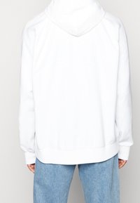 Valkoinen huppari, jossa on pehmeä pinta ja ribattu helma sekä hihat. Selässä on huomaamaton, kohotettu logo. Yhdistetty vaaleansinisiin farkkuihin.