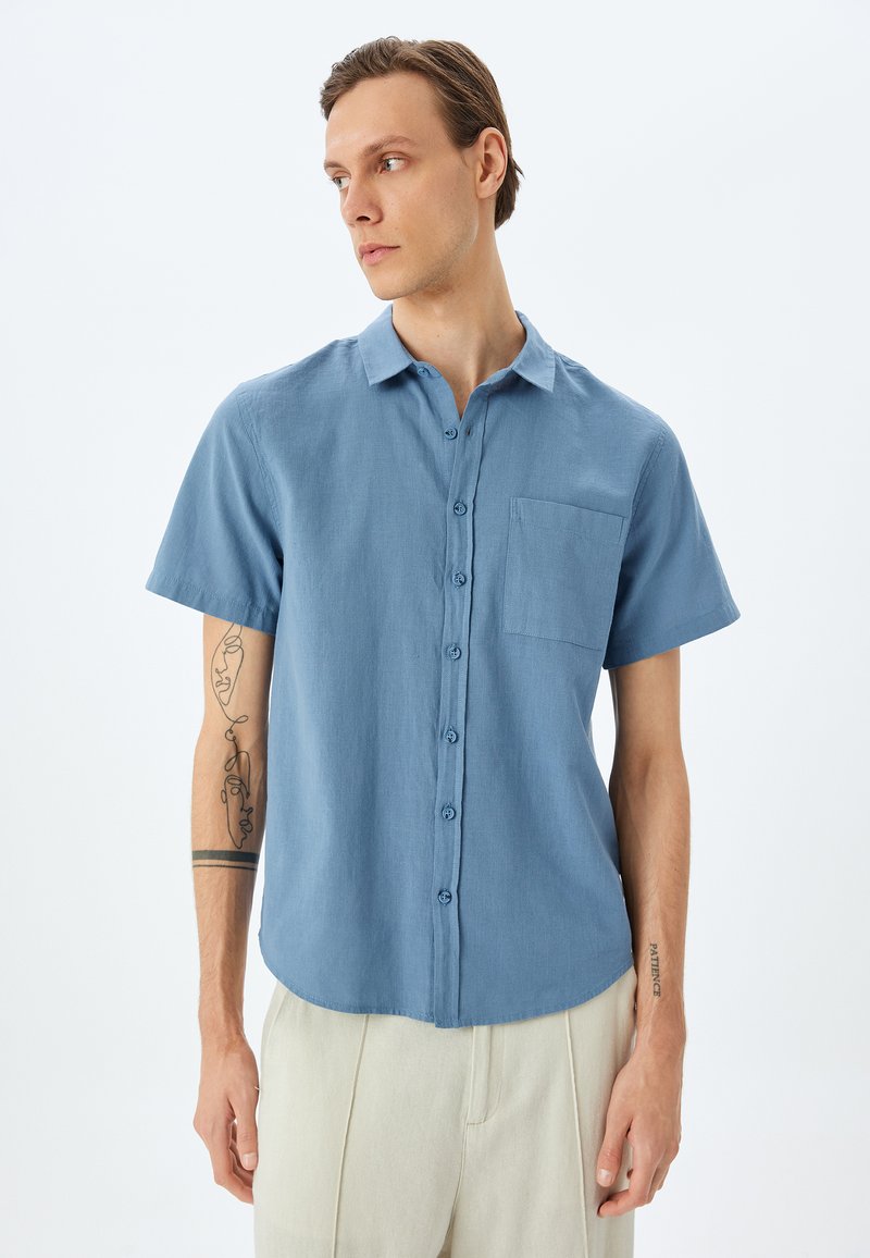 Korte mouwen, knoopsluiting shirt van lichtblauwe stof. Heeft een borstzak, een gebogen zoom en knopen aan de voorkant. Glad textuur.