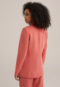 Korallinen blazer, jossa on rakenteellinen muotoilu, kapea istuvuus ja yksittäinen takaventti. Sujuva kangastuntuma ja minimaaliset ommelkorostukset.