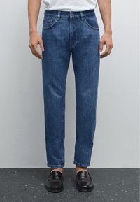 Jean en denim bleu foncé avec une coupe droite, un design à cinq poches et un léger effet fané, porté avec des mocassins noirs sur une surface texturée.