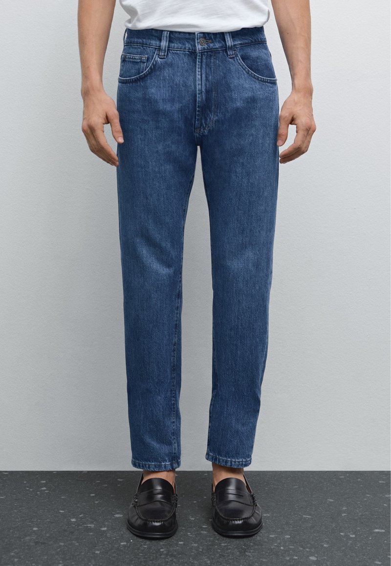 Jean en denim bleu foncé avec une coupe droite, un design à cinq poches et un léger effet fané, porté avec des mocassins noirs sur une surface texturée.