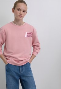 Pink sweatshirt with a crew neckline, featuring a "RACER" logo graphic in white and red. Paired with light blue denim jeans.