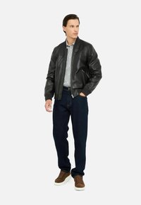Giubbotto bomber in pelle nera con zip frontale, indossato sopra una camicia a righe blu chiaro, abbinato a jeans in denim scuro e scarpe da ginnastica marroni.