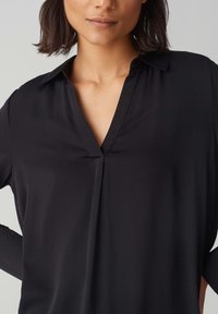 Blusa nera con scollo a V, texture liscia, colletto piegato e maniche lunghe. Presenta una leggera piega sul davanti.