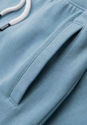 Tela azul claro con un primer plano de un bolsillo lateral vertical y un cordón blanco de una prenda, mostrando la textura de punto y detalles de costura.