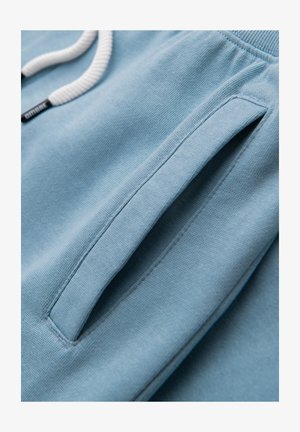 Hellblauer Stoff mit einer Nahaufnahme einer vertikalen Seitentasche und weißem Kordelzug eines Kleidungsstücks, der Strickstruktur und Nähdetails zeigt.