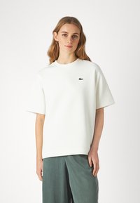 Maglietta oversize in cotone bianco con colletto tondo, maniche corte e un piccolo logo verde sulla parte sinistra del petto. Tessuto morbido.