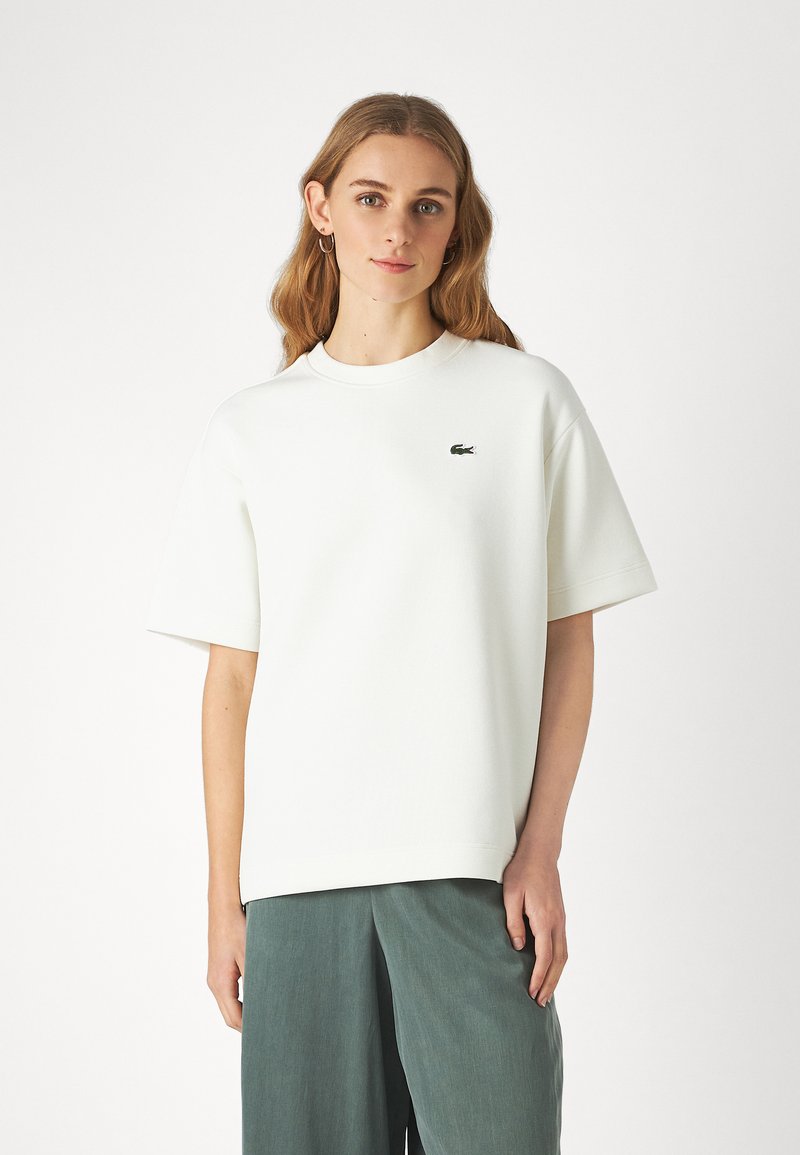 Maglietta oversize in cotone bianco con colletto tondo, maniche corte e un piccolo logo verde sulla parte sinistra del petto. Tessuto morbido.