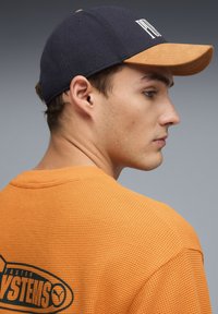 Camisa naranja con textura y gráficos verdes, gorra azul marino con visera color beige, que presenta un logo en la parte frontal. Vista de perfil de una persona.